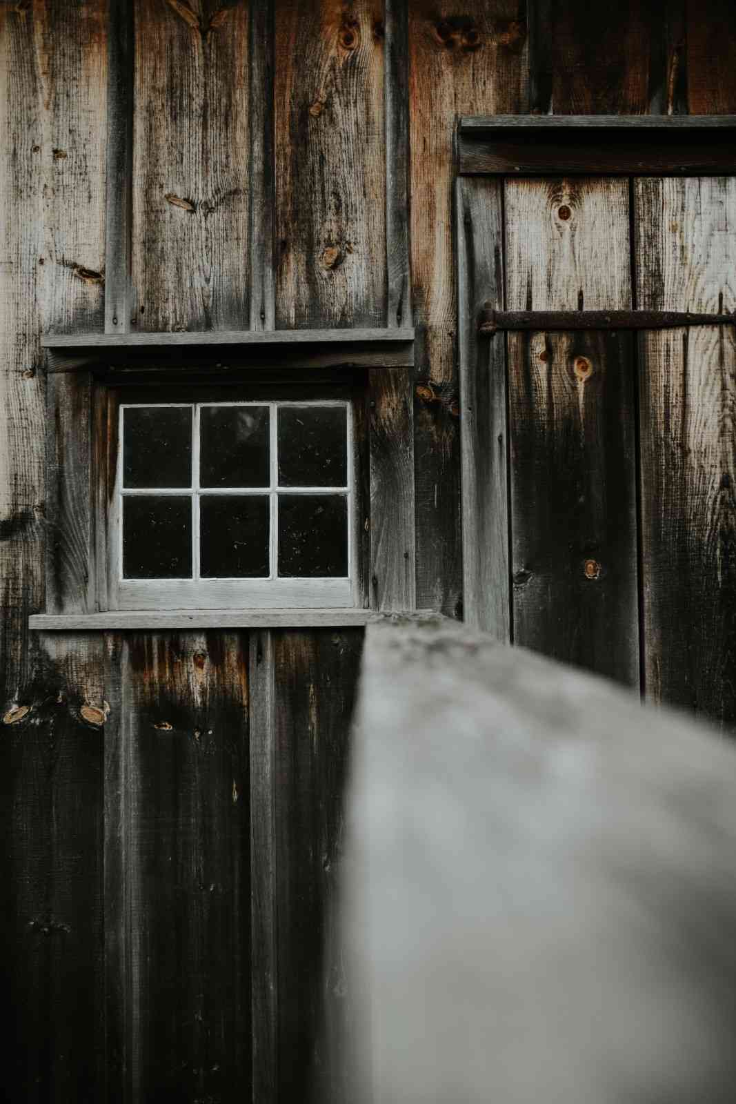 Weathered dark wooden shed exterior with a small white six-pane window and rustic door hinge.