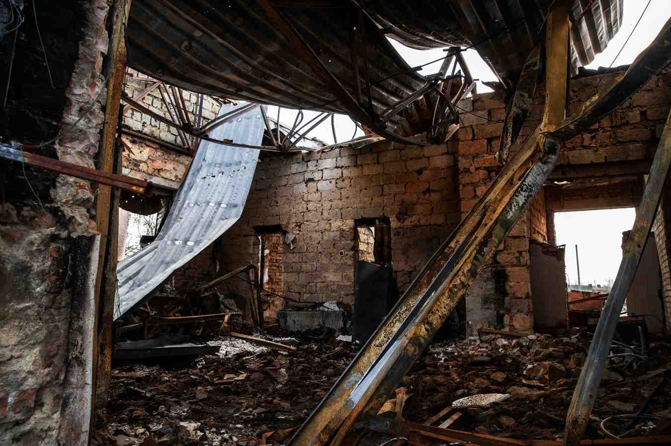 Interior of a destroyed brick building with a collapsed metal roof, burnt beams, and rubble.