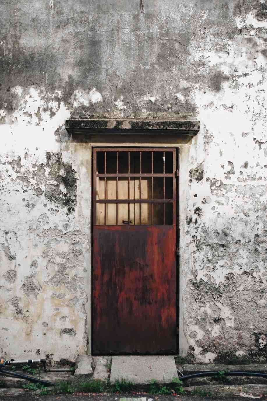 A rusted metal barred door set into a weathered, peeling white concrete wall.