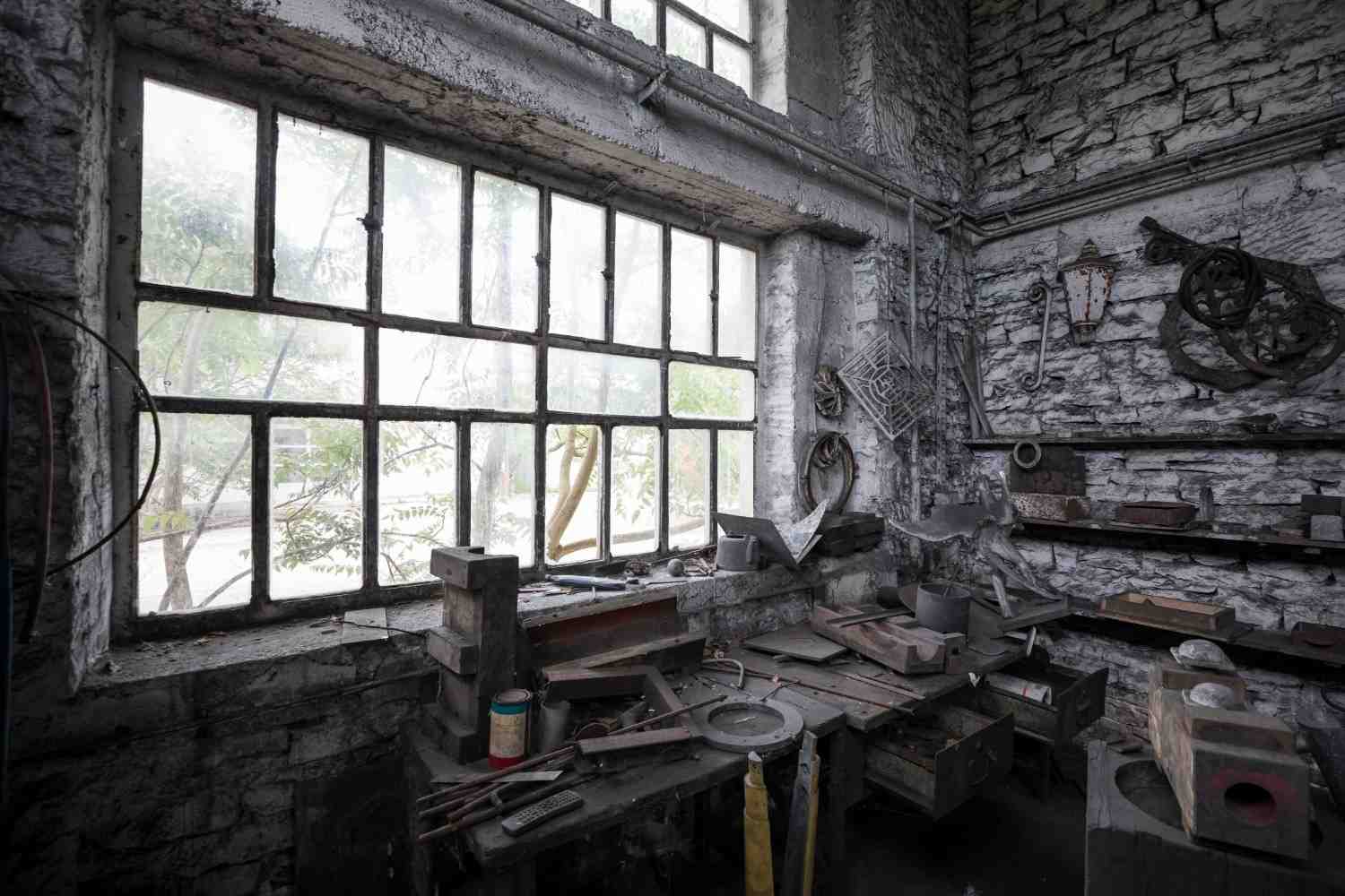 Interior of an abandoned stone workshop with a large window and benches covered in metal debris.
