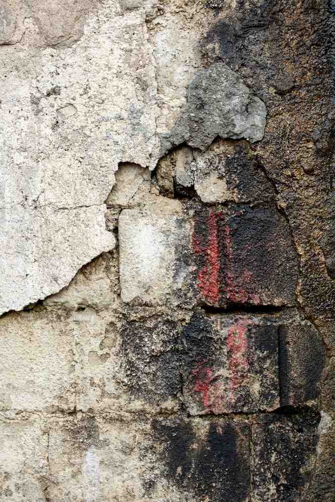 Peeling white wall revealing exposed concrete blocks, heavy dark stains, and red paint marks.