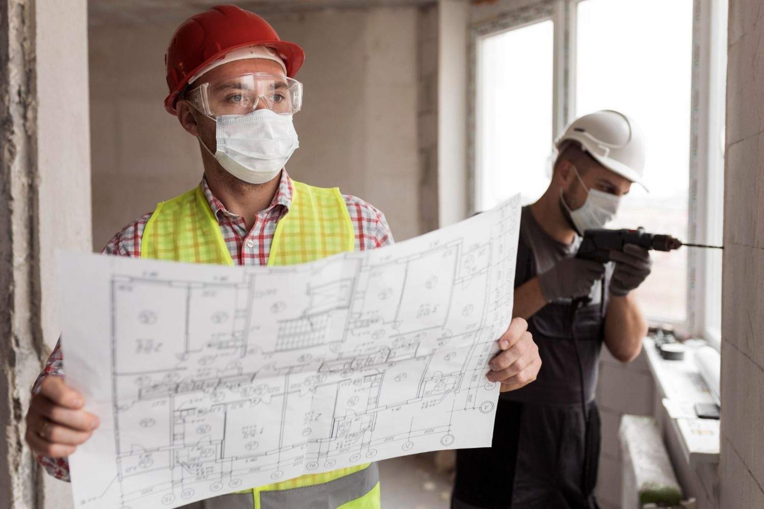 Two construction workers in masks; one holds blueprints while the other drills a wall.