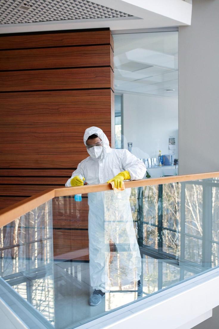 Mold remediation technician in a white hazmat suit cleaning a glass railing with a spray bottle in a modern building.