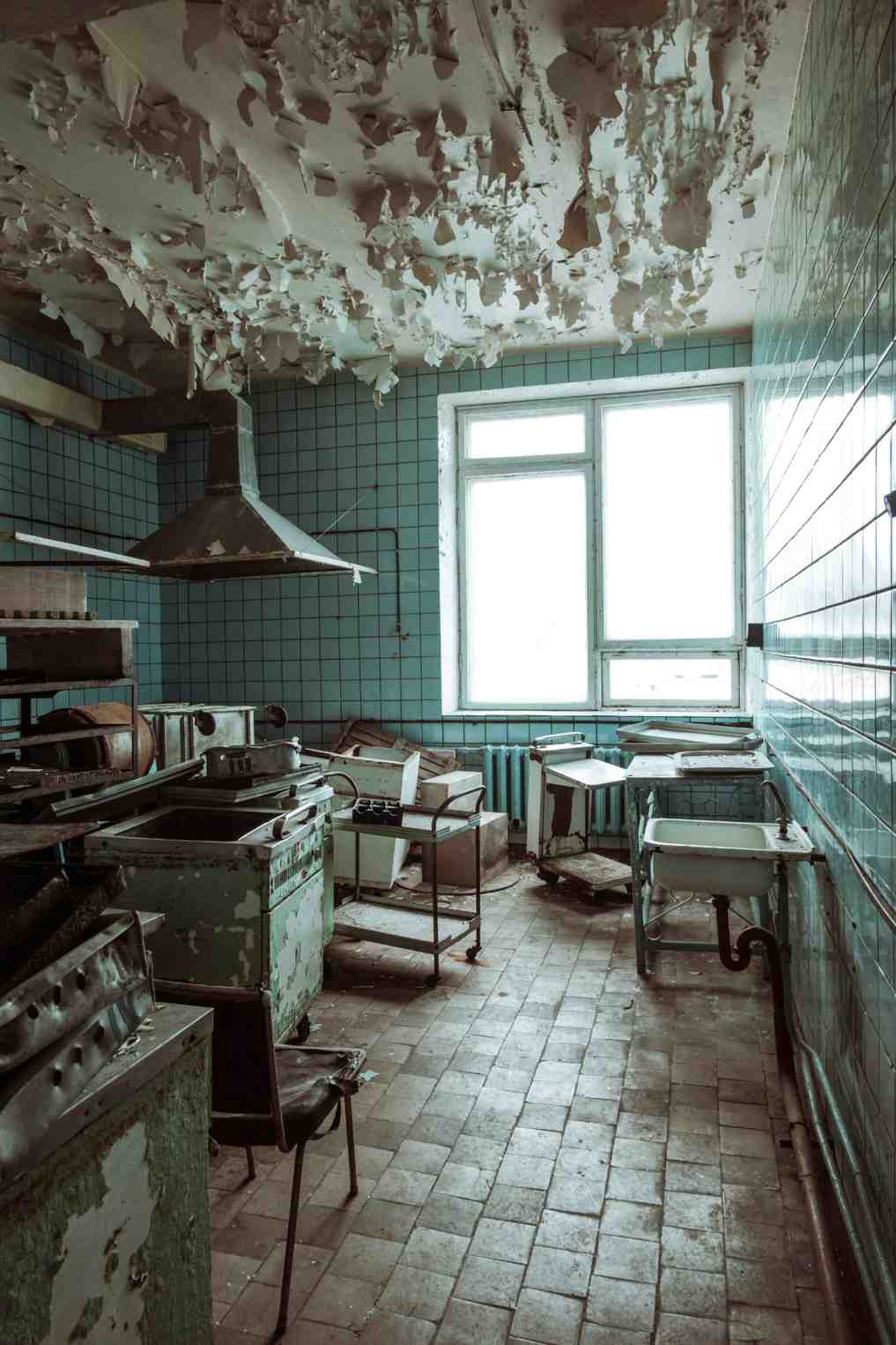 An abandoned commercial kitchen with blue tiled walls and a severely water-damaged ceiling with heavy peeling paint